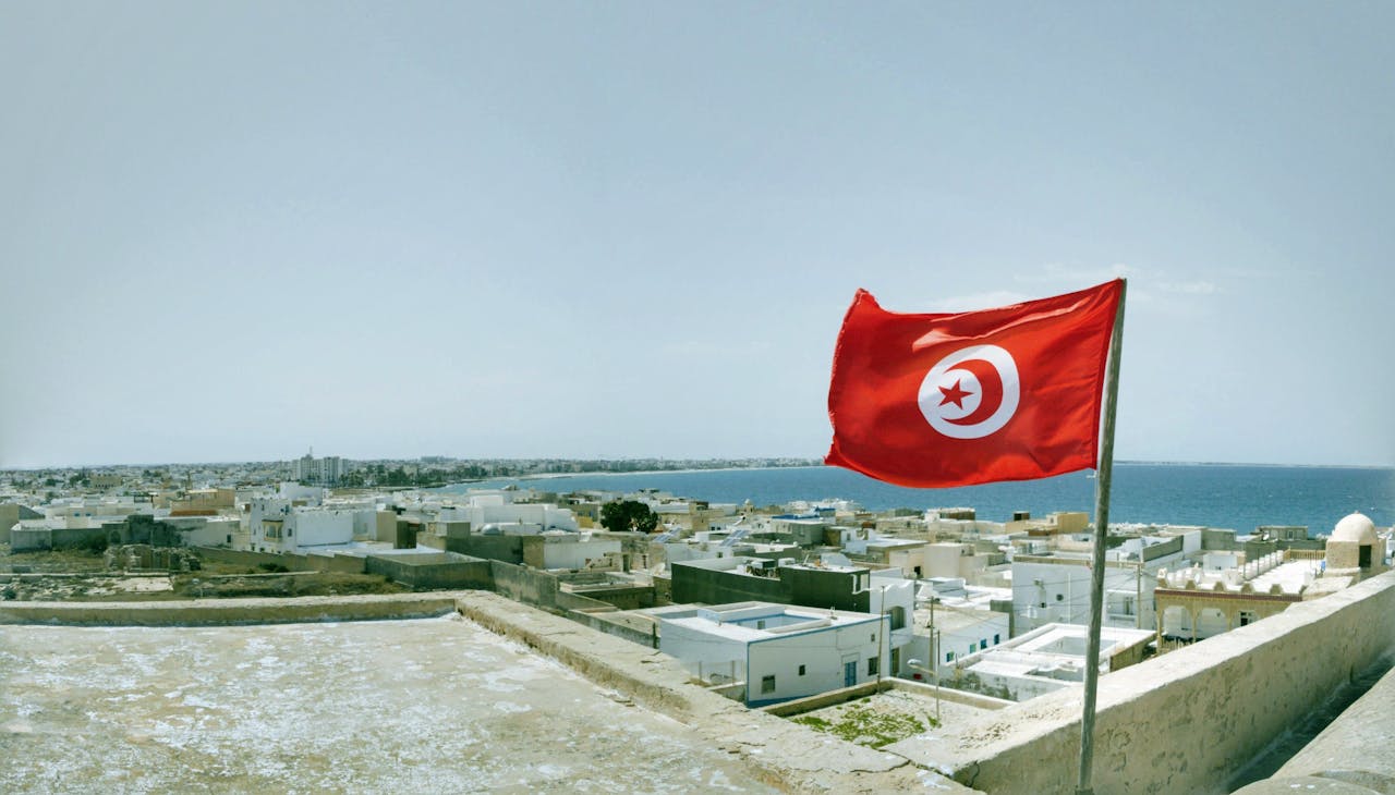 gallery-1 A vibrant Tunisian flag waving over the coastal town of Mahdia with a waterfront view.